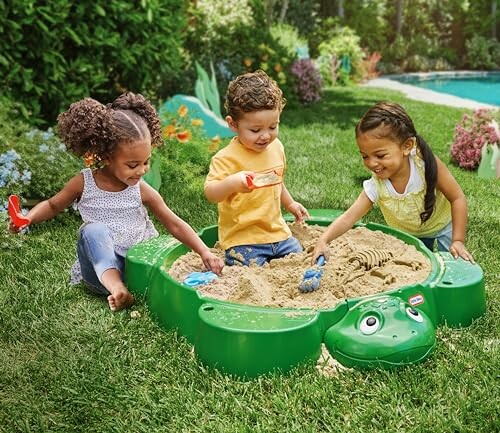 Three children playing in a green sandbox outdoors.