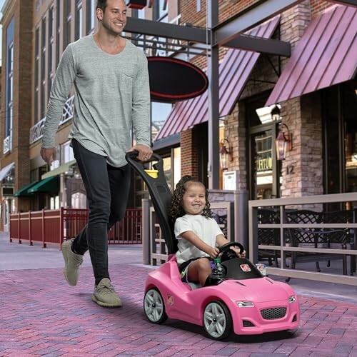 Man pushing a child in a pink toy car on a city street.