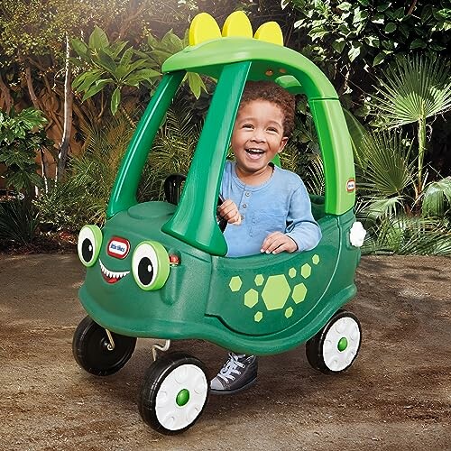 Child smiling in a green dinosaur-themed toy car outdoors.