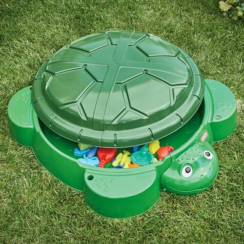 Green turtle-shaped sandbox with lid, filled with colorful toys on grass.